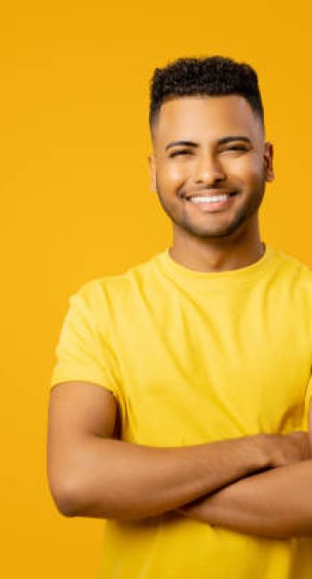 Smiling confident Indian guy wearing casual t-shirt stands with arms crossed isolated on yellow, studio shot of cheerful latin businessman, success concept, copy space